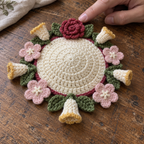 A fingertip beside a rose-red crocheted rose, with cream and gold honeysuckle tubes and pale-pink flowers surrounding an ivory domed coaster.