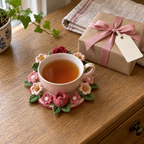 A blush teacup on a pink and red crochet bouquet coaster beside a kraft-wrapped gift with a rose-pink ribbon and blank tag on a wooden sideboard.