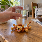 An adult hand sets a glass down onto a golden Lion crochet coaster. Its disc is pressed flat while its open-mouth head, two eyes, amber loop mane and legs rise around the glass.