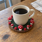 A plain ivory mug of coffee on a dark-green crochet coaster with a brown rope edge and small red-and-white mushrooms around the rim.