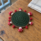 A dark-green crochet coaster without a cup, with a brown rope edge and small red-and-white mushrooms lying outward on a wooden kitchen table.