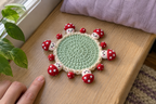 Kawaii Faces crochet mushroom coaster: mint base, cream edge, red caps, white spots, with no cup, the mushrooms lying out round the rim.