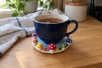 Rainbow Ring crochet mushroom coaster: sky blue base, white edge, red, orange, yellow, green, blue and violet caps, white spots, with a cup set on it, the mushrooms standing up round its foot.