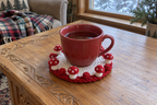 Snowy Christmas crochet mushroom coaster: white base, red edge, red caps, white snow spots, with a cup set on it, the mushrooms standing up round its foot.