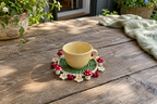 Summer Meadow crochet mushroom coaster: grass green base, white edge, red caps, white spots, four white mini daisies, with a cup set on it, the mushrooms standing up round its foot.