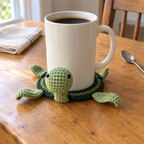A Classic Green crocheted turtle holds an ivory coffee mug on a wooden kitchen table, with its round light-green head standing beside the cup.