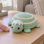 A mint Baby Hatchling crochet turtle with cream shell rings, pink cheeks and a white eggshell cap rests beside a fingertip on a wooden table.