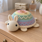 Two gold rings and a pair of small earrings rest on the rounded pastel shell of an Easter Egg crochet turtle with a cream head and cream bunny ears on a bedroom dresser.