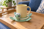 Baby Hatchling pop-up crochet turtle coaster, mint and cream with pink cheeks and a white eggshell cap, shown with a mug set on it, its head standing up beside the cup and its feet raised.