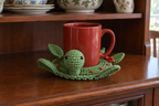 Christmas Wreath pop-up crochet turtle coaster, a green shell with red berries and a little red bow, shown with a mug set on it, its head standing up beside the cup and its feet raised.