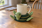 Classic Green Turtle pop-up crochet turtle coaster, dark green with olive ribs and a light-green face and feet, shown with a mug set on it, its head standing up beside the cup and its feet raised.