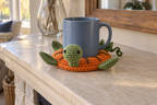 Pumpkin pop-up crochet turtle coaster, orange segments with a green leaf and curly vine beside the tail, shown with a mug set on it, its head standing up beside the cup and its feet raised.