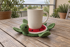 Watermelon pop-up crochet turtle coaster, red fruit, a white inner rind and a green edge, shown with a mug set on it, its head standing up beside the cup and its feet raised.