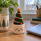 A crochet Fir Tree with a gold star, cream garland and red baubles in a cream smiling pot on a wooden desk beside a closed laptop and a mug.