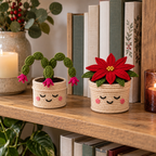 Crochet Christmas Cactus and Poinsettia plants in cream smiling pots on a wooden shelf beside books and candles.