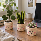 Crochet Monstera, snake plant and pink-tipped succulent in cream smiling pots on a wooden home office desk beside a laptop and mug.