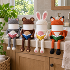 Cat, Bear, Bunny and Fox crochet reading roll holders sit on a bathroom vanity with their hats on, open pink, blue, yellow and green books and hanging legs.