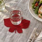 A maple-red Red Maple crochet coaster under a clear water glass on a linen-covered lunch table beside a plate and fork.