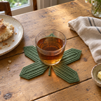A sage-green star-shaped Sweetgum crochet coaster under a glass of tea on a wooden breakfast table beside bread and a linen napkin.