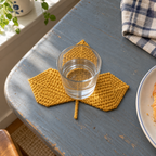 A mustard-gold three-lobed Sycamore crochet coaster under a water glass on a blue-grey wooden table beside a checked towel and plate.
