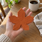 An adult open hand holding a finished pumpkin-orange Sugar Maple crochet coaster above a wooden kitchen table beside a coffee mug.