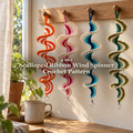 Four crochet ribbon wind spinners in orange and white, pink and ivory, blue and cream, and green and oatmeal hang from a wooden peg rail beside a sunny window, with a mug and plants below and the product title across the centre.