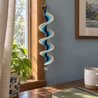 A navy, teal and aqua Lagoon Arch Lace ribbon wind spinner with open ivory arches and a clear bead hangs from a window-frame hook above a wooden desk.