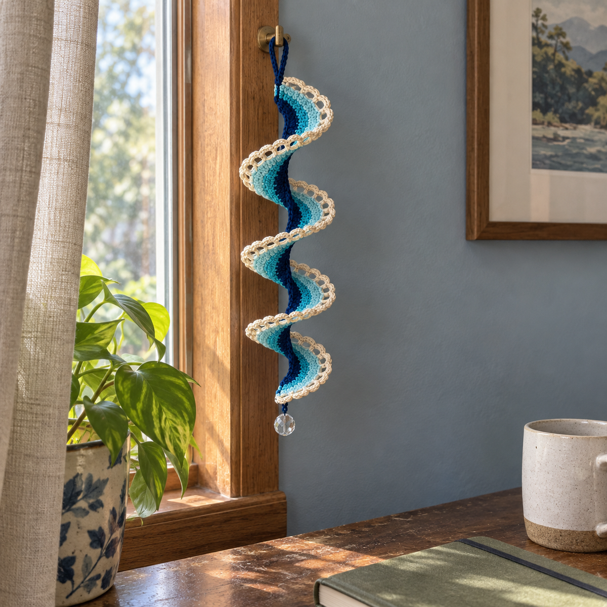 A navy, teal and aqua Lagoon Arch Lace ribbon wind spinner with open ivory arches and a clear bead hangs from a window-frame hook above a wooden desk.