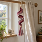 A burgundy, dusty rose and mauve crochet spiral with a cream edge and matching tassel hanging from a wooden curtain rod beside a linen curtain.