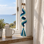 A navy, teal and aqua crochet spiral with cream scallops and a matching tassel hanging in a coastal window overlooking the sea.