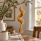 A golden crochet spiral with an ivory open ladder edge and a gold and ivory tassel hanging from a leafy tree branch in warm window light.