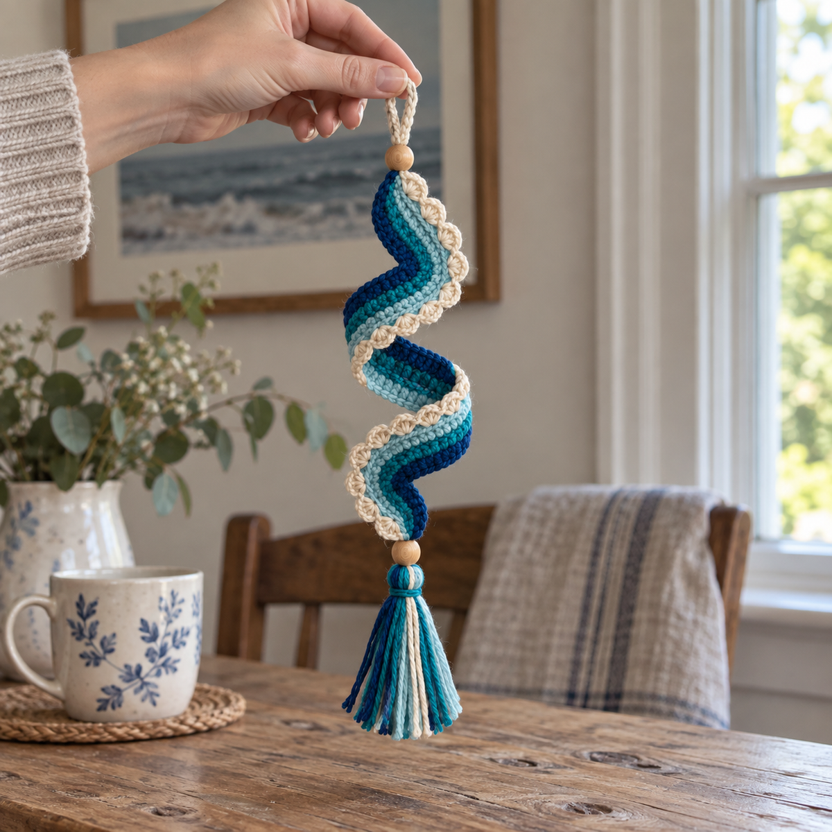 An adult hand holding a navy, teal and aqua crochet spiral ornament by its cream hanging loop in a home by a window.
