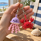 A close view of lilac jellyfish and red crab crochet charms held beside a seashell, showing yarn stitches and embroidered faces.