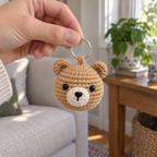 An adult hand holds a silver ring with a small tan crochet bear charm hanging below it in a living room.