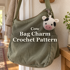 A white cow crochet bag charm with black patches and a pink muzzle hangs from the zip of a sage-green canvas bag in a sunlit hallway, with the title Cow Bag Charm Crochet Pattern across the centre.
