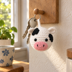 A white cow crochet bag charm hangs beside two house keys from a wooden peg near a sunny kitchen window.