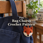 Rust and cream crochet fox head bag charm hanging from the zipper of a navy canvas bag on a wooden chair beside a window, with the title Fox Bag Charm Crochet Pattern.