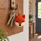 Rust and cream crochet fox head charm hanging beside two house keys from a silver ring on a brass hook mounted on an oak hallway board.
