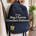 A green crochet frog charm with cream eye bumps hangs from the lower pocket zip of a navy backpack on a wooden hallway peg, with the title Frog / Bag Charm / Crochet Pattern across the centre.