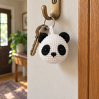 A white and black crochet panda charm hangs beside two house keys from a brass hook in a sunlit entryway.