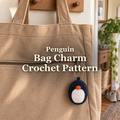 Navy crochet penguin bag charm with a white belly and orange beak hanging from a tan canvas tote zipper in a sunlit entryway, with the title Penguin Bag Charm Crochet Pattern.