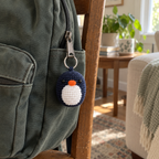 Navy crochet penguin bag charm hanging from the zipper of a green canvas backpack beside a wooden chair in a living room.