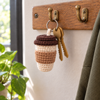 Cream crochet coffee-cup charm with a kraft tan sleeve and brown two-tier lid hanging from a silver keyring with two brass house keys on a brass hook on a wooden key rail beside a window.
