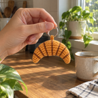 An adult hand holding the silver ring of a golden crocheted croissant charm beside a kitchen window.