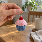 An adult hand holds a blue and pink crochet cupcake by its silver ring above a wooden table beside a ceramic mug and folded linen cloth.