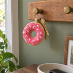 A tan crochet donut with pink glaze and white sprinkles hanging beside two brass house keys on a wooden wall rail.