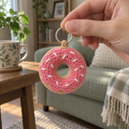 An adult hand holding a pink-glazed crochet donut with white sprinkles by its silver ring in front of a sage sofa.
