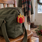 Red and gold crocheted gift box charm hanging from the zipper of an olive canvas bag on a wooden chair.