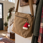 A small red and off-white crochet Santa hat charm hangs from the zipper of a tan canvas bag in a home entryway with Christmas greenery.