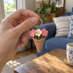 An adult hand holding the silver ring of a small pink and cream crochet bouquet keychain, with a blue sofa and wooden table behind.